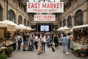 Panoramica del mercato coperto "East Market Presents Pet Market" affollato di persone e cani in un grande edificio industriale con pareti in mattoni a vista.
